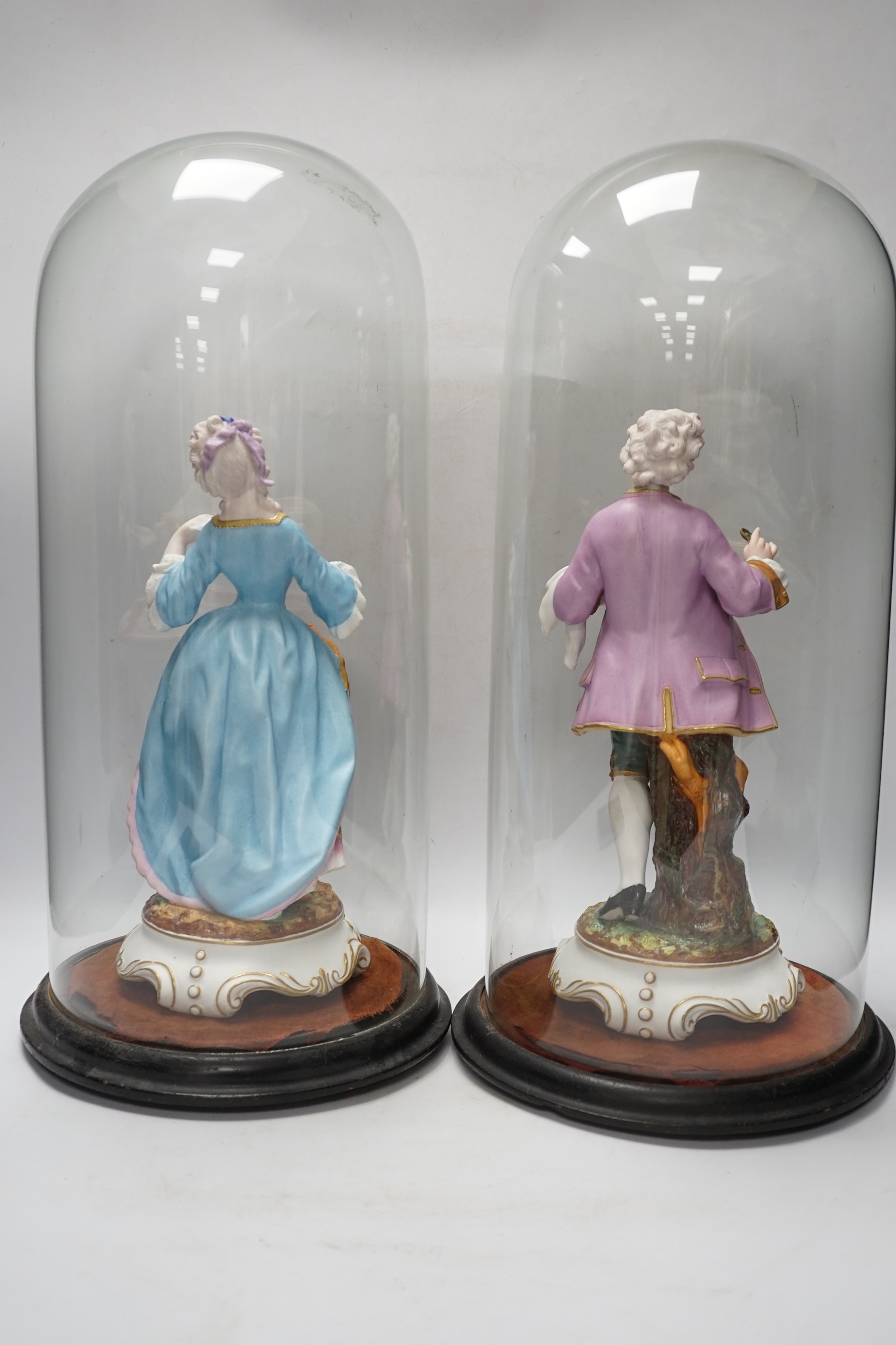 A pair of continental bisque porcelain figures, under glass domes, 48cm overall